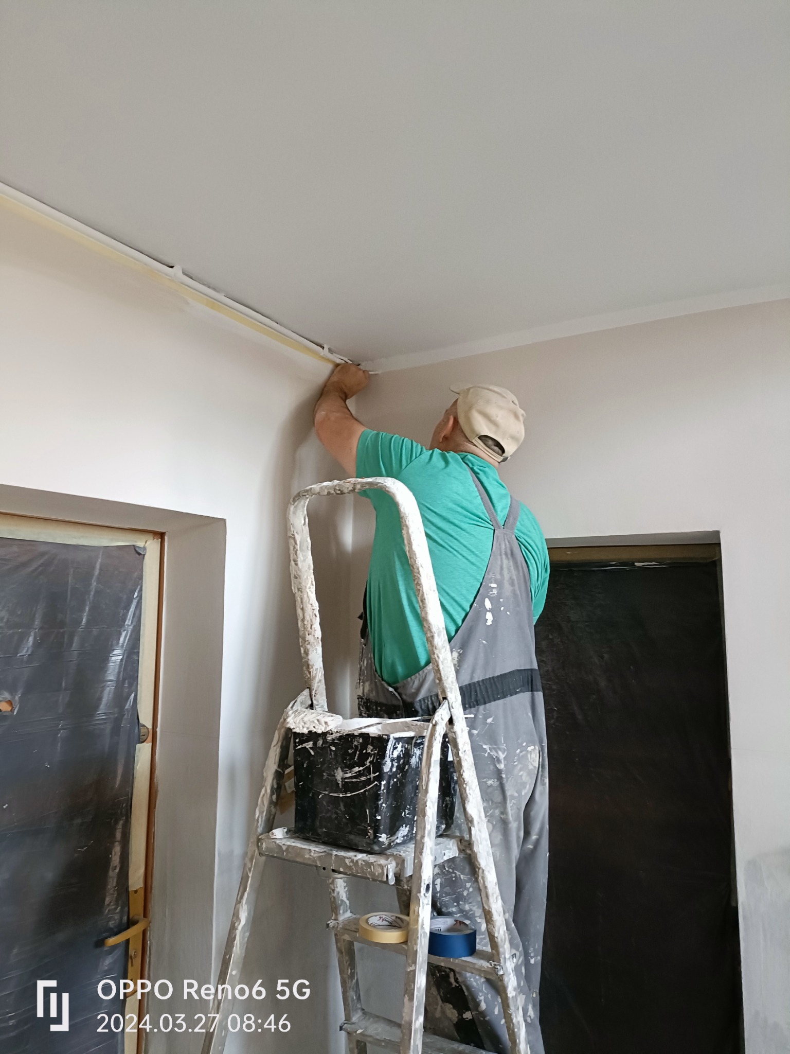 Pracownik na drabinie zabezpiecza taśmą malarską narożnik sufitu w pomieszczeniu przygotowanym do malowania, widoczny zakryty folią otwór drzwiowy.