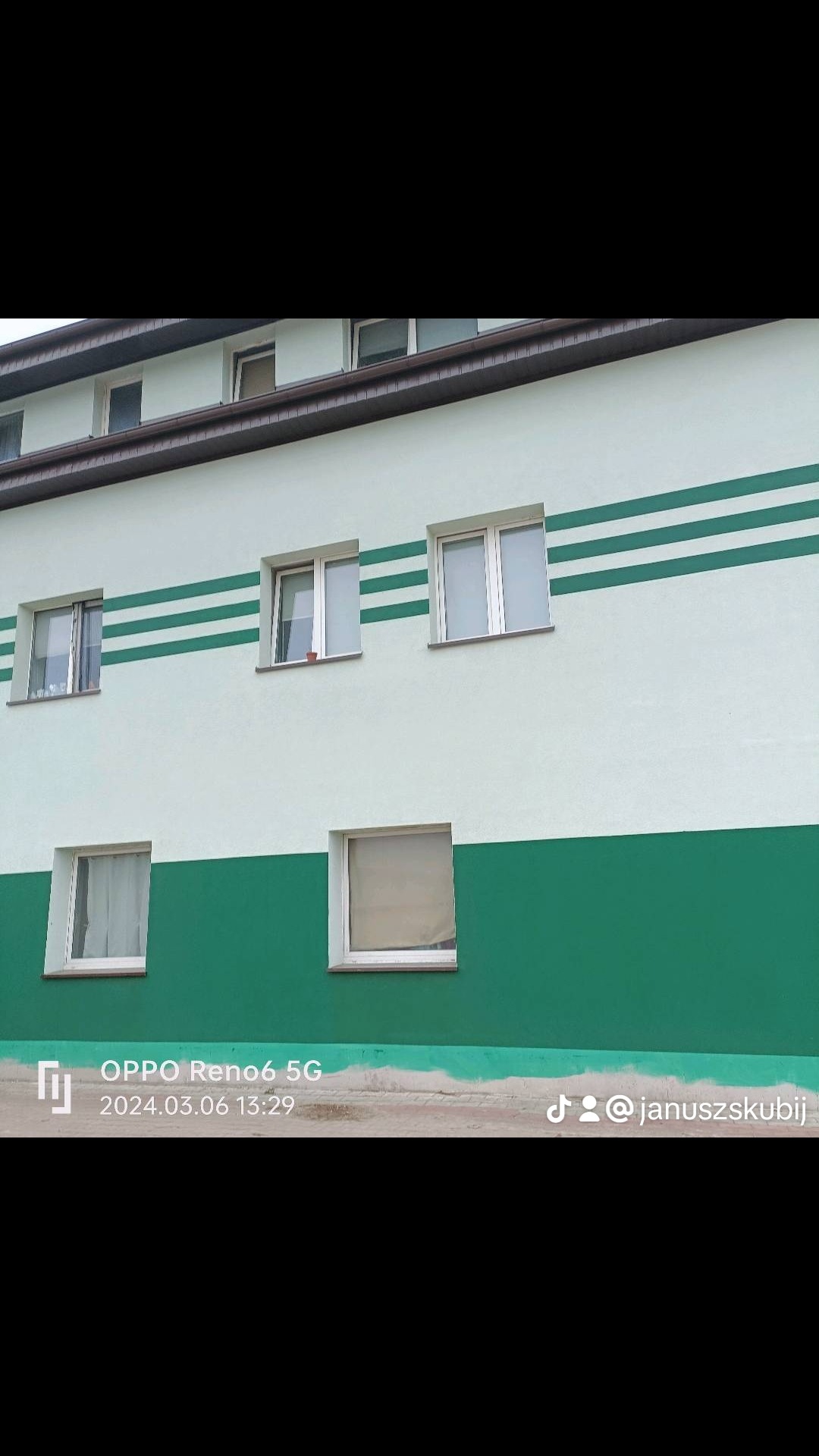 Odnowiona elewacja budynku z białymi ścianami, zielonym pasem na dole i trzema zielonymi paskami w górnej części, okna w białych ramach.