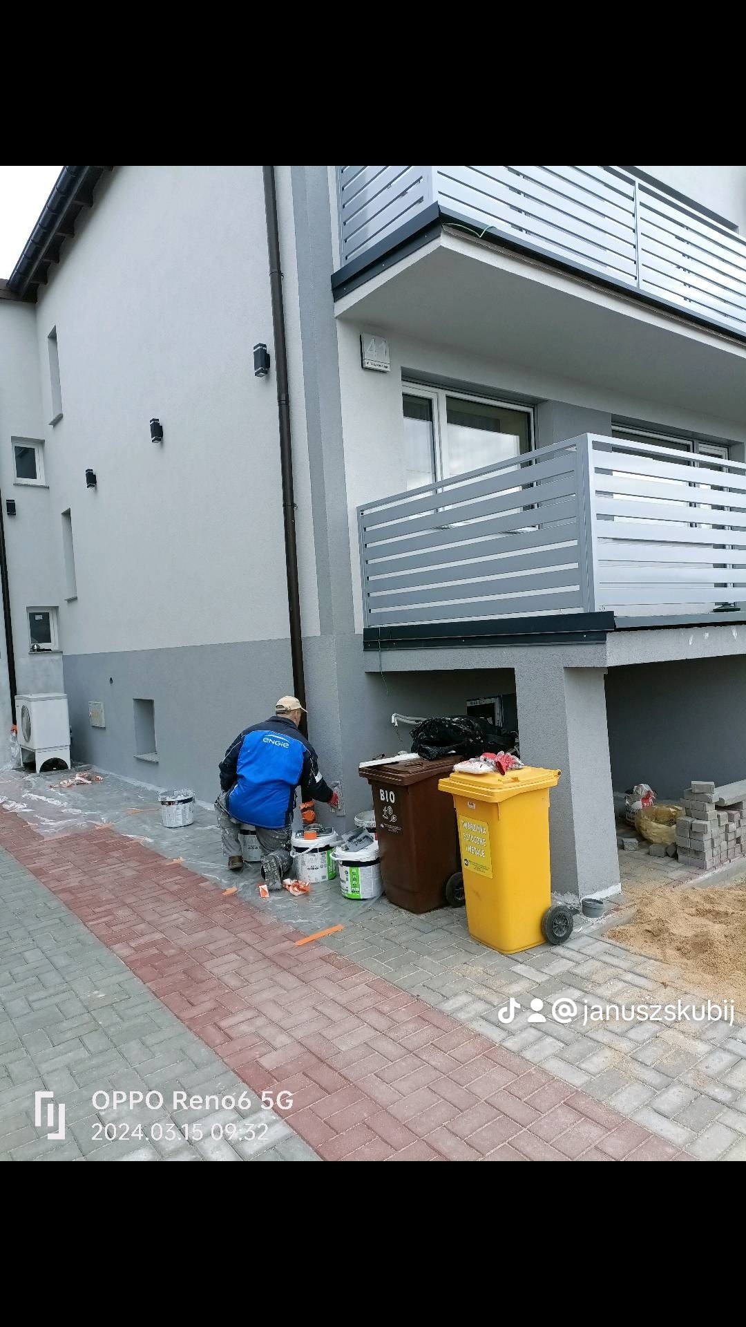 Pracownik w niebieskiej kurtce maluje szarą elewację domu jednorodzinnego, widoczne puszki z farbą, dwie plastikowe beczki na śmieci i częściowo zabezpieczona kostka brukowa.