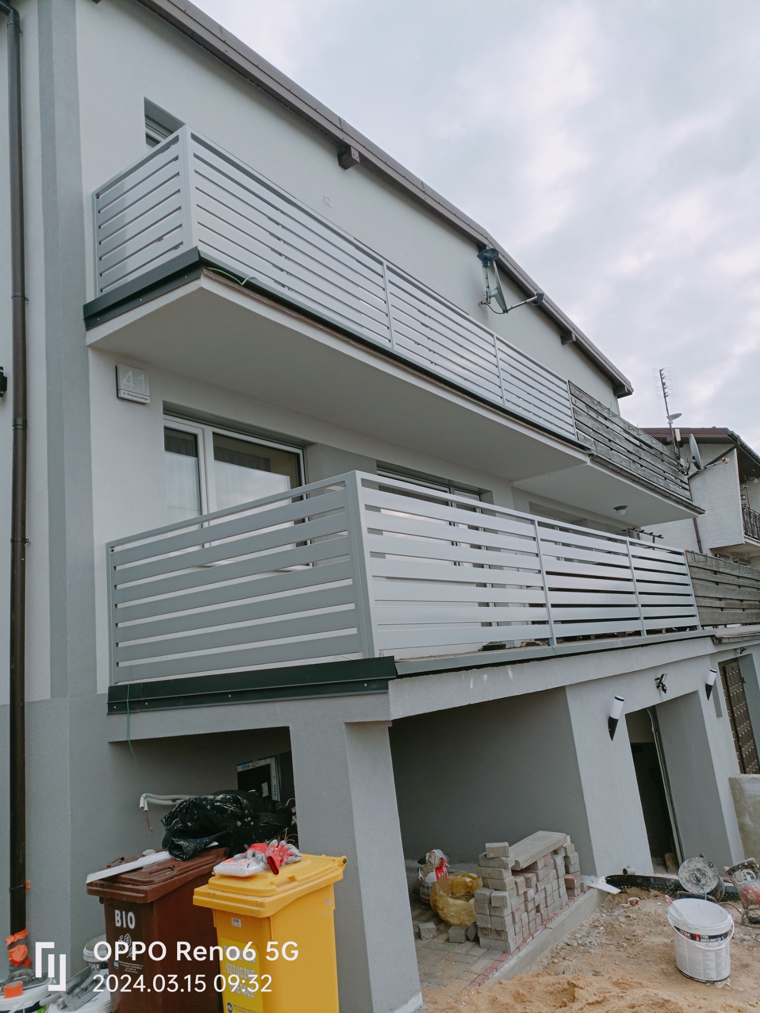 Dwupiętrowy budynek z szarą elewacją i nowoczesnymi, poziomymi balustradami balkonowymi w kolorze szarym; widoczne ślady prac budowlanych na parterze, w tym pojemniki na odpady i materiały budowlane.