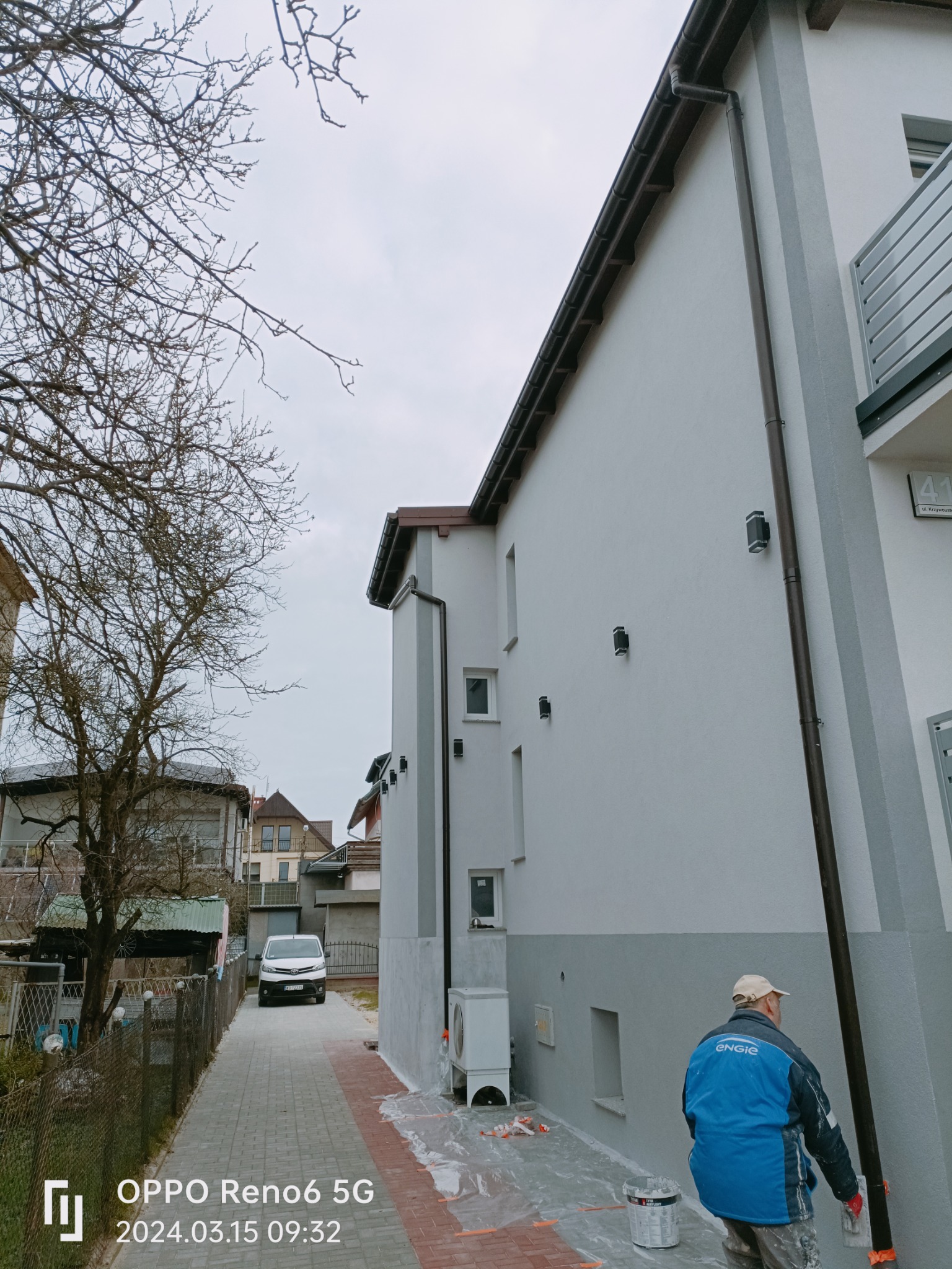 Pracownik w niebieskiej kurtce szpachluje elewację budynku na szaro, widoczne wiadro z farbą, zabezpieczona folią kostka brukowa i fragment podwórka z samochodem w tle.