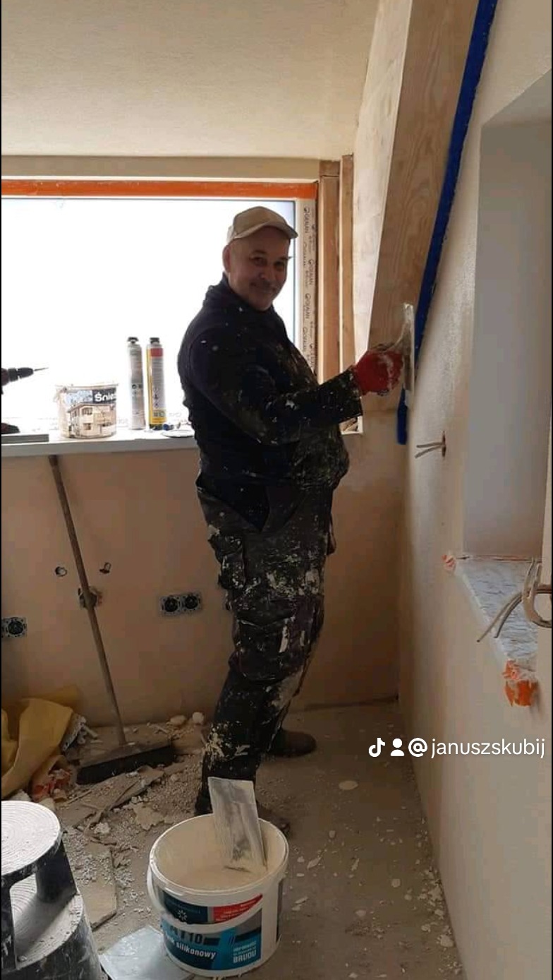 Mężczyzna w roboczym ubraniu, z uśmiechem, nakłada szpachlę na ścianę w trakcie prac wykończeniowych, obok stoi wiadro ze szpachlą.