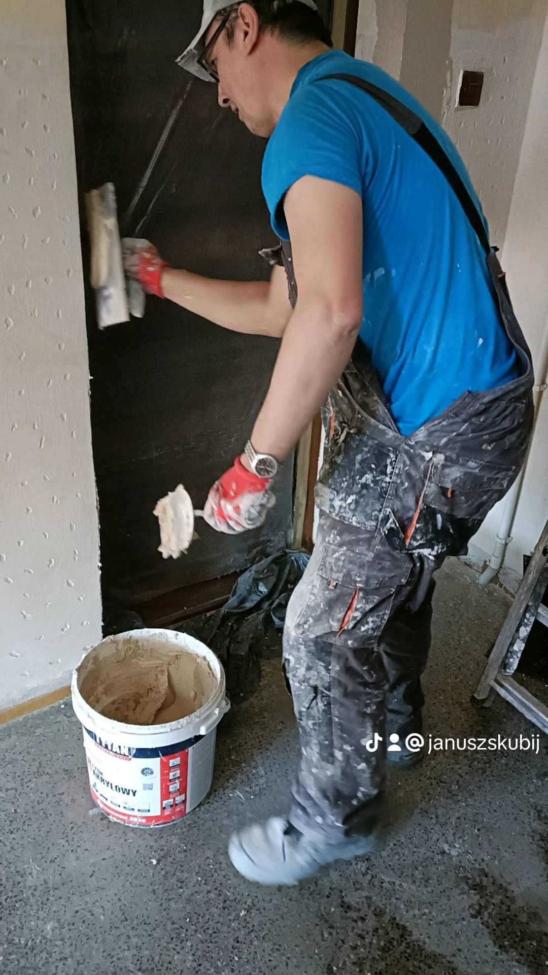 Mężczyzna w okularach i roboczym ubraniu nanosi szpachlę na ścianę, używając szpachelki; obok stoi wiadro z masą szpachlową.