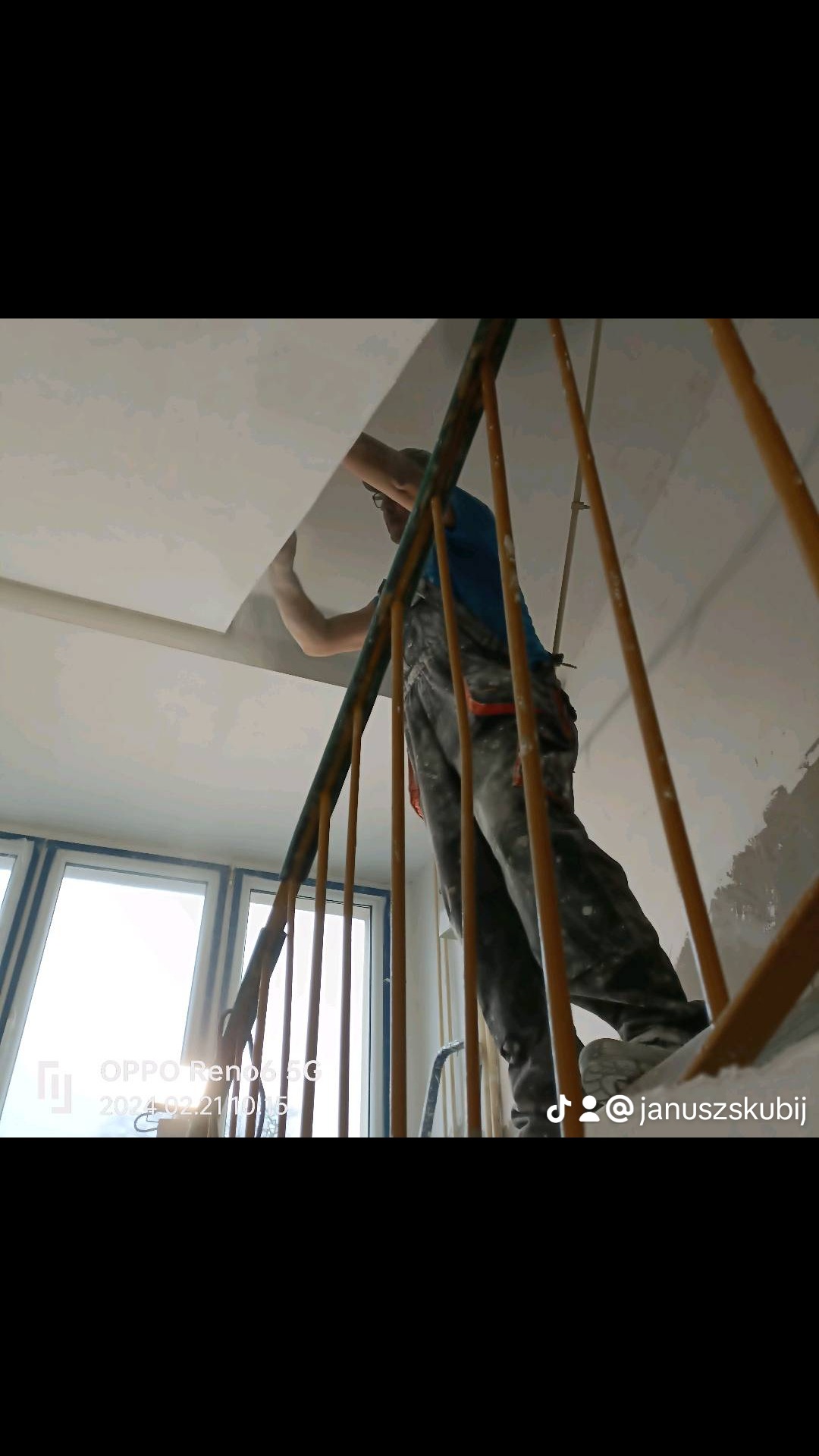 Pracownik w kombinezonie, stojąc na schodach, wygładza świeżo nałożoną warstwę gładzi na suficie, widok z dołu przez metalową barierkę schodów.