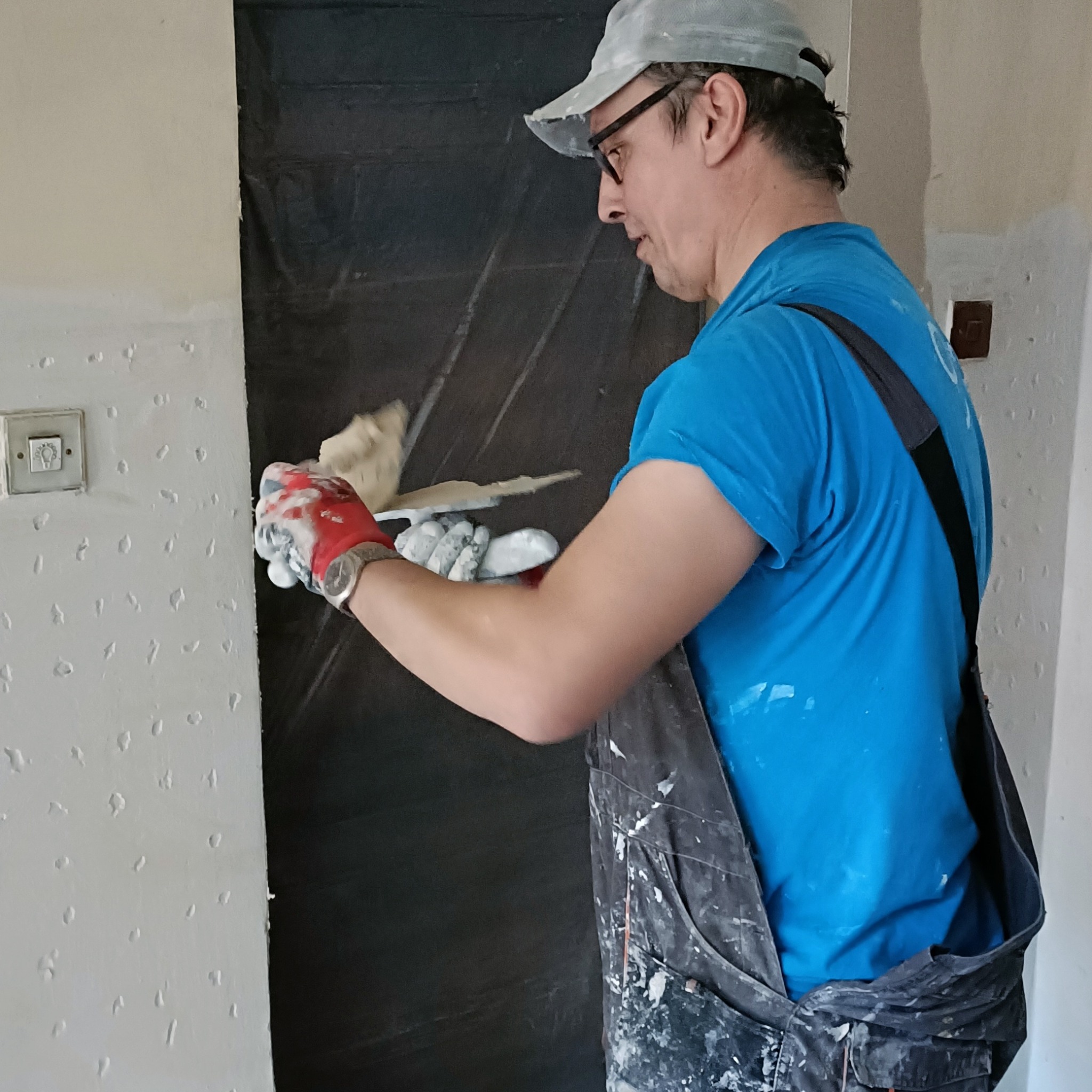 Mężczyzna w okularach i roboczym ubraniu, w trakcie nakładania szpachli na ścianę z widoczną fakturą, obok zabezpieczonych folią drzwi.
