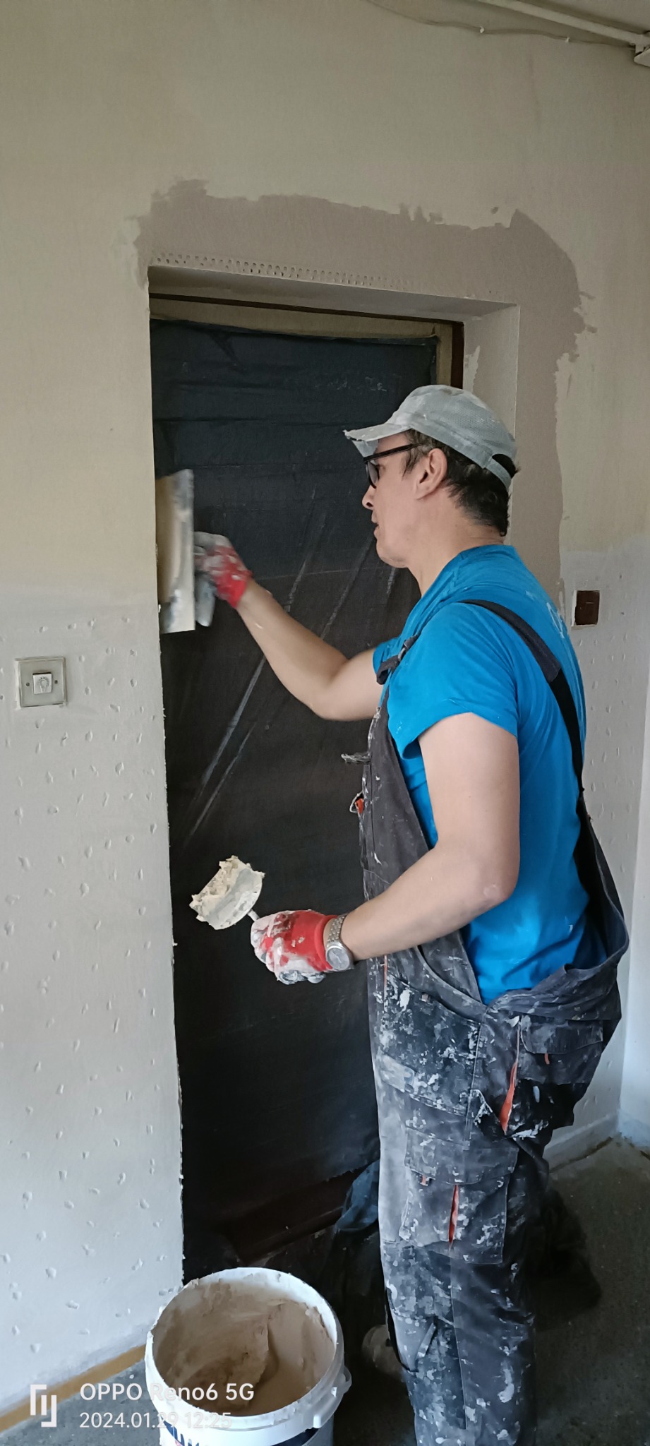 Pracownik w okularach i czapce, w kombinezonie roboczym, nakłada masę szpachlową na ścianę przy futrynie drzwi, używając szpachelki; widoczny kubeł z masą i ślady po nakładaniu na ścianie.