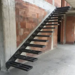 Surowe, metalowe schody bez poręczy, zamontowane na ścianie z cegły i betonu w surowym stanie, widok z dołu na wznoszące się stopnie.