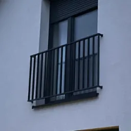 Czarna, metalowa balustrada balkonowa z pionowymi szczeblami, zamontowana na elewacji budynku z szarą roletą okienną.