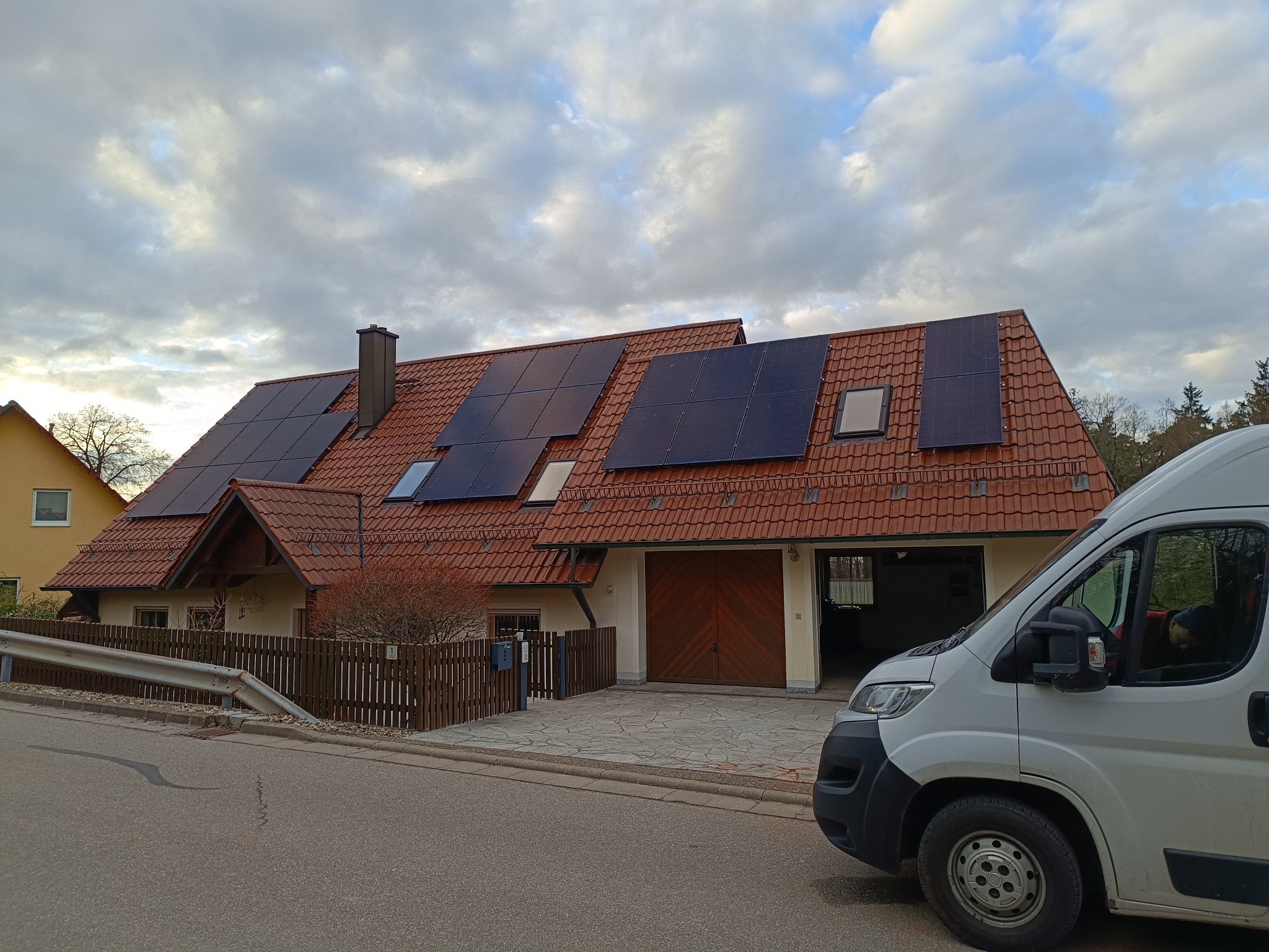 Dom jednorodzinny z panelami słonecznymi na dachu z czerwonej dachówki, zaparkowany biały samochód dostawczy z boku, pochmurne niebo.