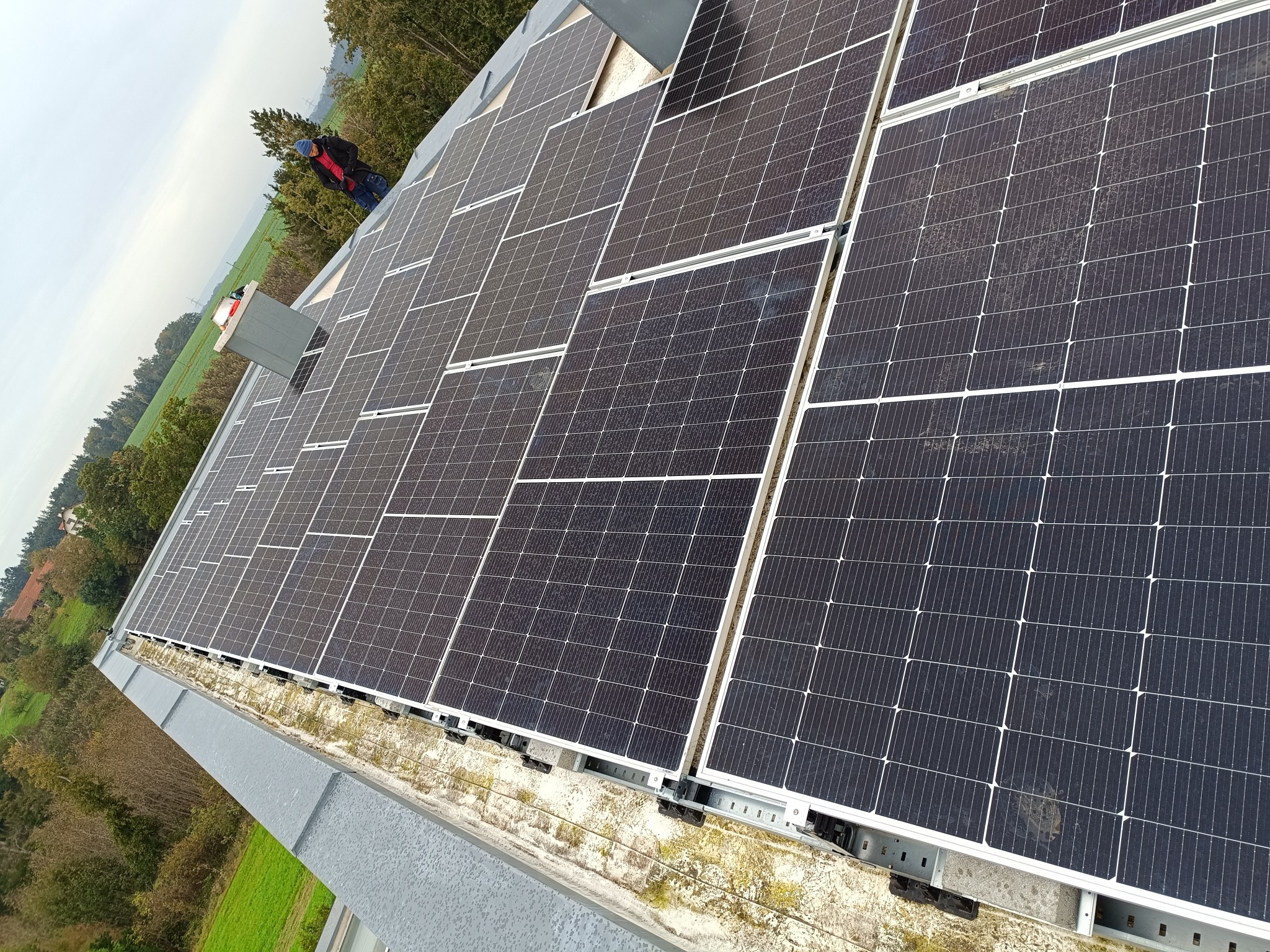 Widok z góry na dużą instalację paneli słonecznych na płaskim dachu, z pracownikiem w oddali sprawdzającym system, otoczoną drzewami i zielenią.