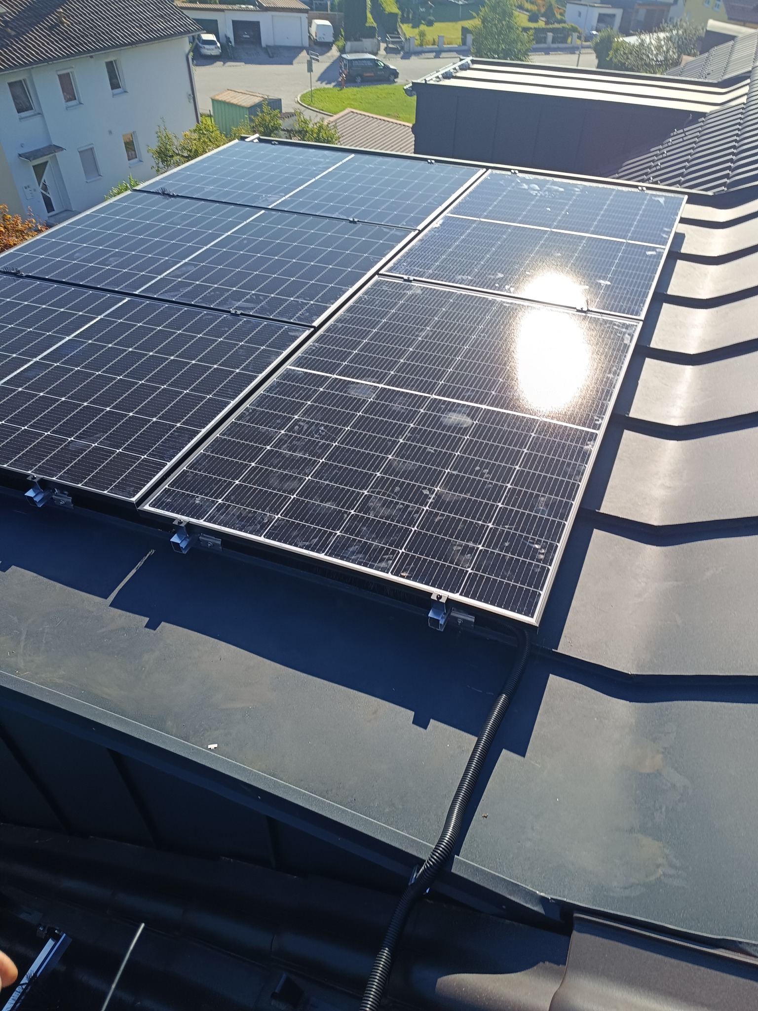 Panel słoneczny na dachu z blachy trapezowej, widoczne mocowania i peszel z przewodami, w tle budynki mieszkalne.