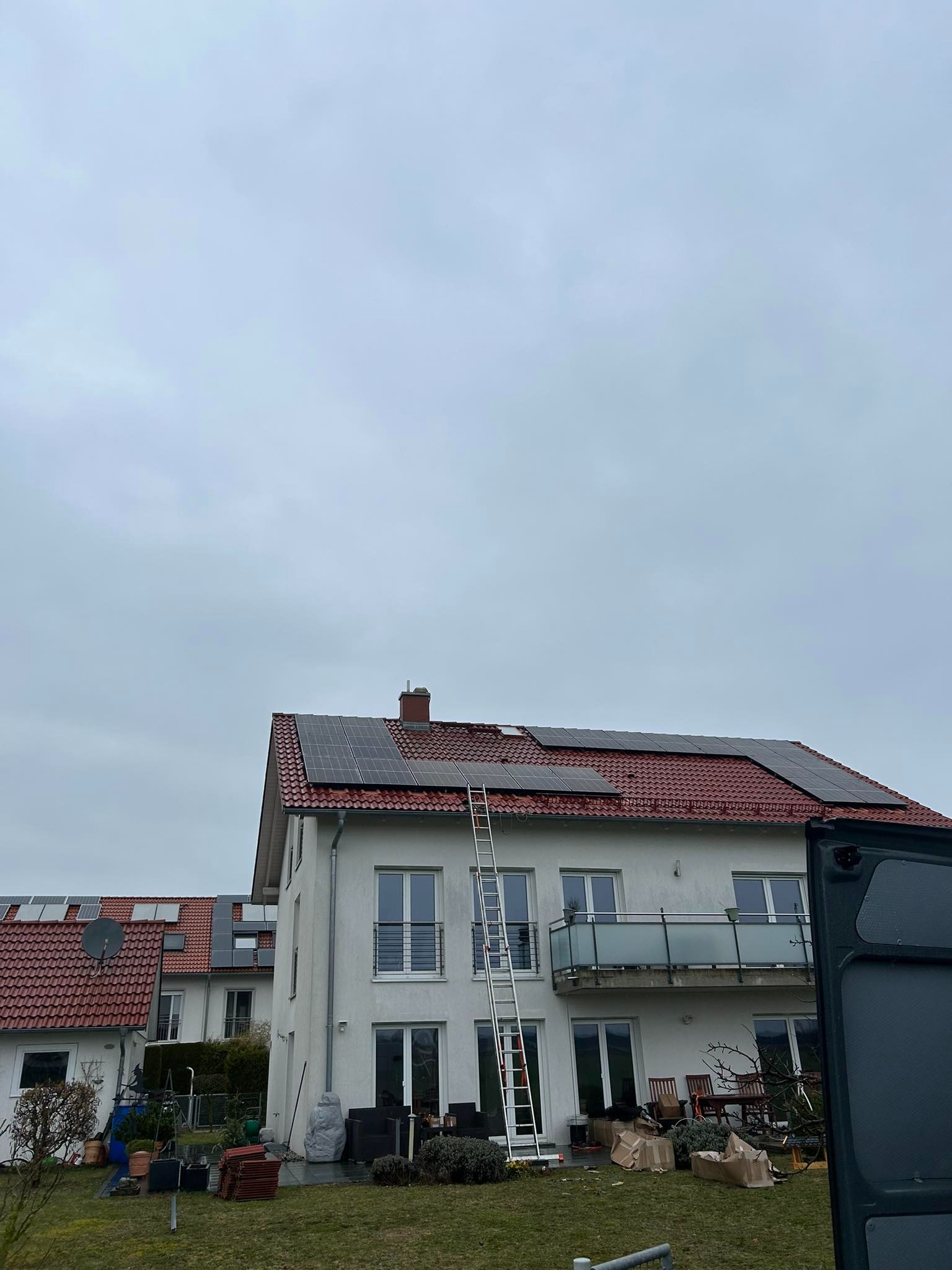 Montaż paneli na dachu domu jednorodzinnego z czerwoną dachówką, drabina oparta o dach, pochmurne niebo.