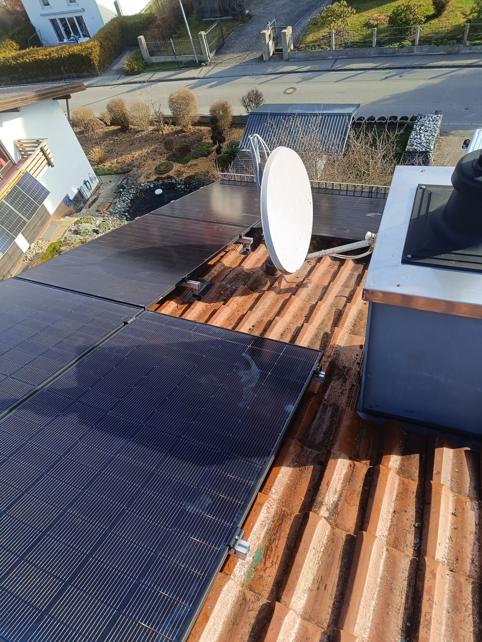 Montaż paneli na dachu z czerwonej dachówki, widoczna antena satelitarna i fragment komina, w tle dom i zadbany ogród.