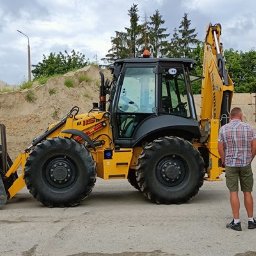 EDU-Consulting SZKOLENIA I DORADZTWO Anna Wiechowska, Danuta Namiak - spółka cywilna - Żółta koparko-ładowarka New Holland 115B z dwoma mężczyznami w tle, jeden w kamizelce odblaskowej i kasku, na tle pryzmy piasku i betonowego muru.