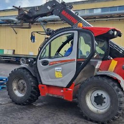 EDU-Consulting SZKOLENIA I DORADZTWO Anna Wiechowska, Danuta Namiak - spółka cywilna - Czerwono-szary wózek widłowy Manitou na zewnątrz, obok sterty drewnianych palet, z widocznym operatorem w kabinie.