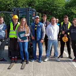 EDU-Consulting SZKOLENIA I DORADZTWO Anna Wiechowska, Danuta Namiak - spółka cywilna - Grupa osób, w tym jedna kobieta trzymająca dyplom, pozuje na tle wózka widłowego i stosu rur, prawdopodobnie po ukończonym kursie lub szkoleniu.