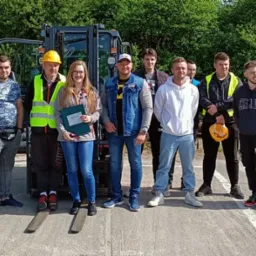 Grupa osób, w tym jedna kobieta trzymająca dyplom, pozuje na tle wózka widłowego i stosu rur, prawdopodobnie po ukończonym kursie lub szkoleniu.