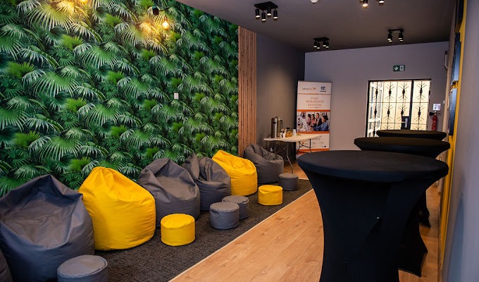 Nowoczesna przestrzeń biurowa z pufami w odcieniach szarości i żółci na tle ściany z tropikalnym motywem liści, stoliki koktajlowe w czarnych pokrowcach, plakat informacyjny w tle.