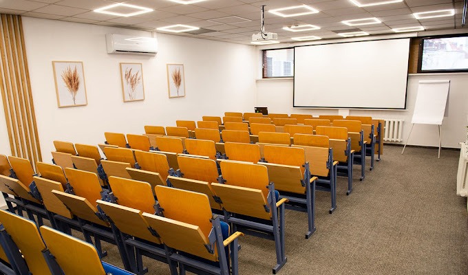 Sala konferencyjna z rzędami krzeseł z drewnianymi oparciami, projektor sufitowy, biały ekran, flipchart i dekoracje ścienne z motywem roślinnym.