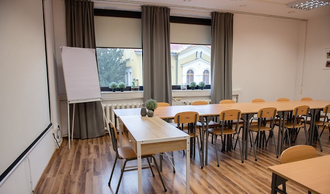 Sala konferencyjna z długim stołem, krzesłami, flipchartem i oknami z widokiem na budynek