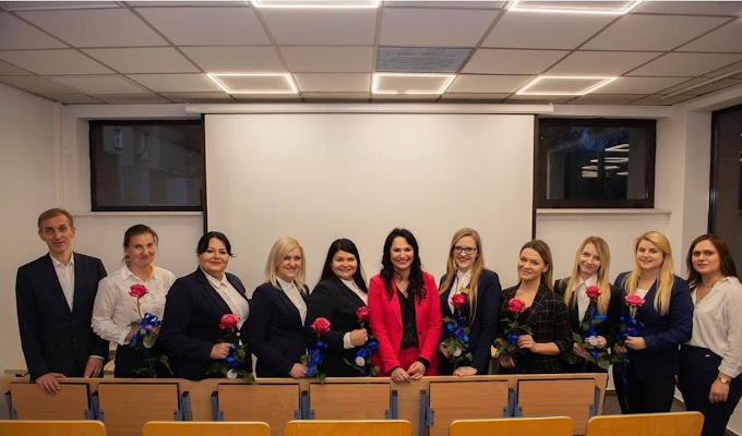 Grupa osób w garniturach, trzymających róże, stojąca na tle sali konferencyjnej z projektorem i krzesłami, prawdopodobnie po ukończonym szkoleniu.