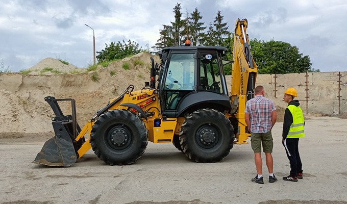 Żółta koparko-ładowarka New Holland 115B z dwoma mężczyznami w tle, jeden w kamizelce odblaskowej i kasku, na tle pryzmy piasku i betonowego muru.