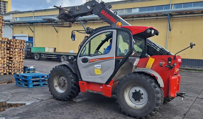 Czerwono-szary wózek widłowy Manitou na zewnątrz, obok sterty drewnianych palet, z widocznym operatorem w kabinie.