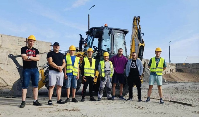 Grupa ośmiu pracowników budowlanych w żółtych kaskach i kamizelkach odblaskowych pozuje na tle żółtej koparko-ładowarki na placu budowy z betonowymi blokami w tle.