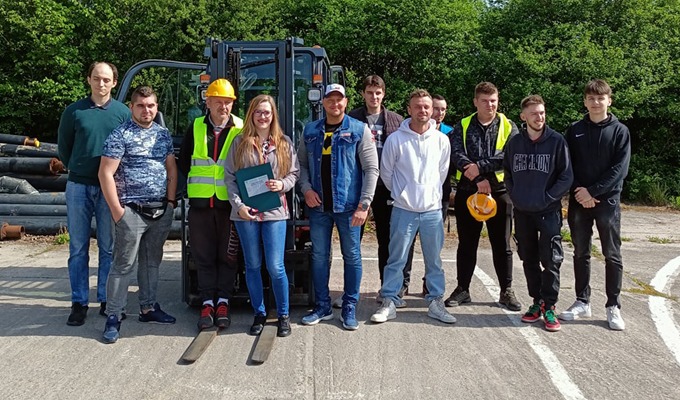 Grupa osób, w tym jedna kobieta trzymająca dyplom, pozuje na tle wózka widłowego i stosu rur, prawdopodobnie po ukończonym kursie lub szkoleniu.