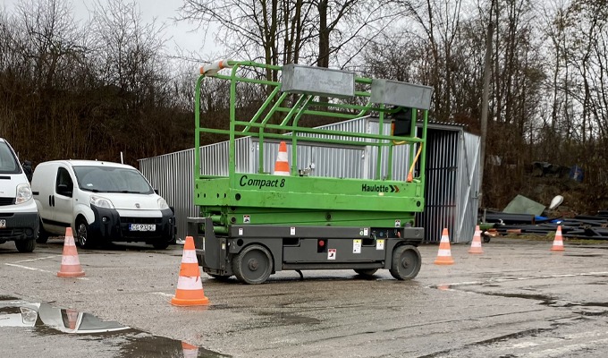 Zielony podnośnik nożycowy Compact 8 Haulotte na mokrym placu, otoczony pachołkami i samochodami dostawczymi w tle.