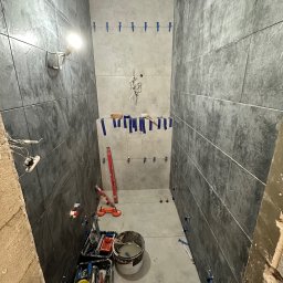 WallCraft-Remonty - Wąska łazienka w trakcie układania ciemnych, grafitowych płytek na ścianach, widoczne narzędzia glazurnicze, poziomica, wiadro z zaprawą oraz niepodłączone kable elektryczne.