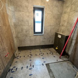 WallCraft-Remonty - Łazienka w trakcie prac: Ułożone płytki ścienne w odcieniach szarości, częściowo ułożone płytki podłogowe z niebieskimi krzyżykami dystansowymi, widoczna instalacja podtynkowa WC, pion poziomicy...