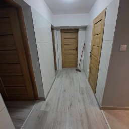 WallCraft-Remonty - Korytarz po remoncie z panelami podłogowymi imitującymi drewno, wykończony płytkami imitującymi marmur na ścianach, z trzema drewnianymi drzwiami, widoczna czerwona zmiotka oparta o ścianę.