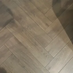 Ułożona pod kątem brązowa podłoga panelowa w jodełkę, widoczna niebieska ściana z białą listwą przypodłogową.