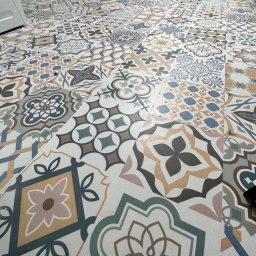 WallCraft-Remonty - Wzór podłogi z płytek ceramicznych o różnorodnych, geometrycznych i kwiatowych motywach w odcieniach beżu, brązu, szarości i błękitu, ułożonych na jasnej spoinie, widoczne narzędzia malarskie w tle.