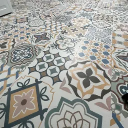 Wzór podłogi z płytek ceramicznych o różnorodnych, geometrycznych i kwiatowych motywach w odcieniach beżu, brązu, szarości i błękitu, ułożonych na jasnej spoinie, widoczne narzędzia malarskie w tle.