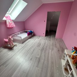 WallCraft-Remonty - Pomalowany na różowo pokój dziecięcy na poddaszu z szarymi panelami podłogowymi, łóżkiem, konikiem na biegunach i oknem dachowym z wiszącą sukienką.