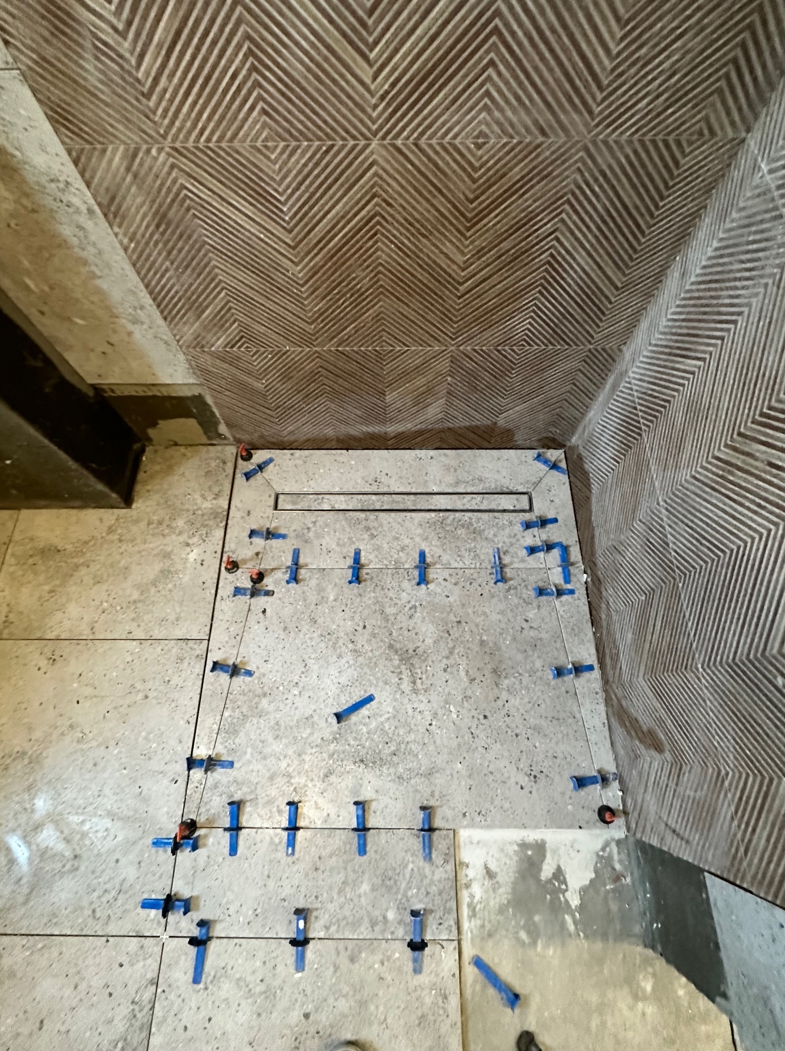 Fragment łazienki w trakcie układania płytek podłogowych i ściennych. Widoczne niebieskie klipsy poziomujące, odpływ liniowy oraz wzór na ścianie w jodełkę.