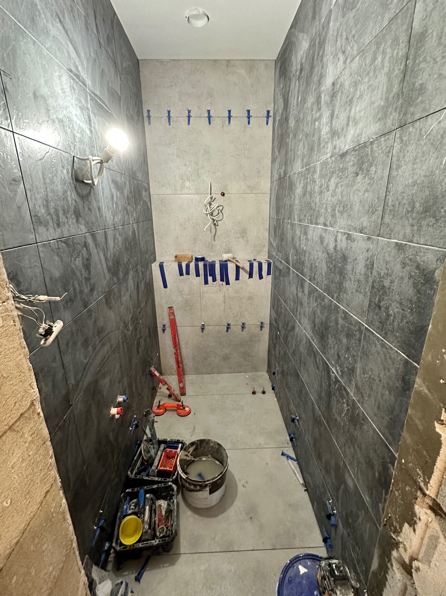 Wąska łazienka w trakcie układania ciemnych, grafitowych płytek na ścianach, widoczne narzędzia glazurnicze, poziomica, wiadro z zaprawą oraz niepodłączone kable elektryczne.