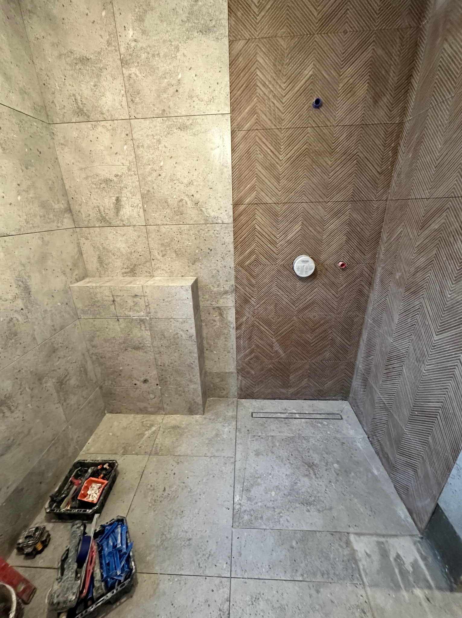 Wnętrze kabiny prysznicowej z wyłożonymi płytkami ścianami: jedna ściana z płytek imitujących kamień, druga z wzorem w jodełkę, widoczne narzędzia glazurnicze na podłodze.