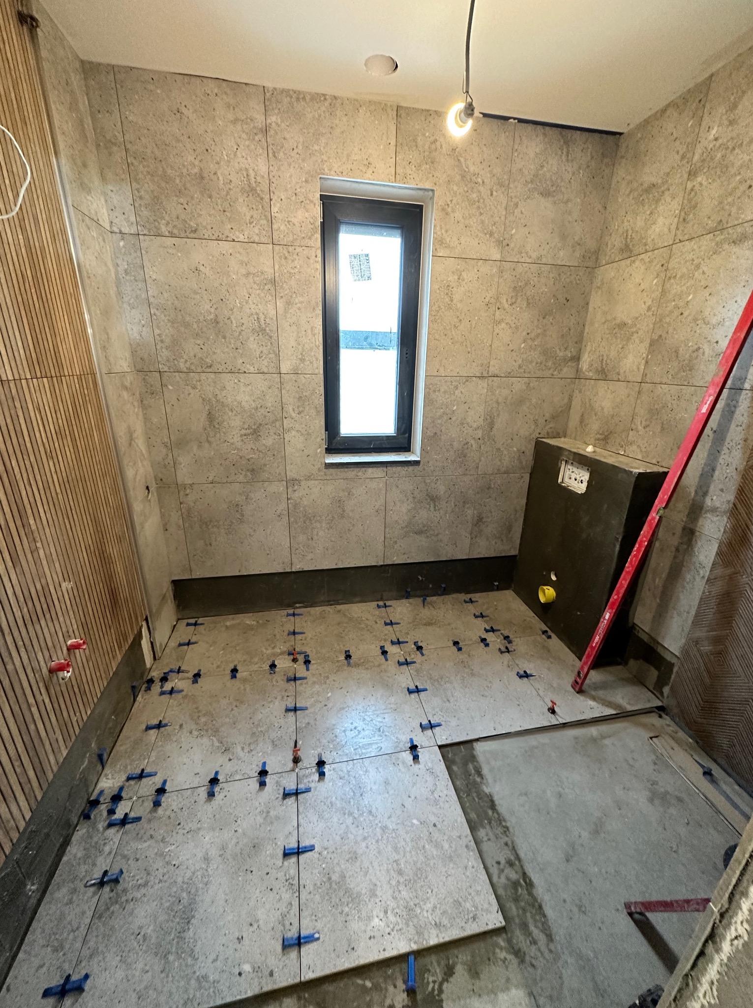 Łazienka w trakcie prac: Ułożone płytki ścienne w odcieniach szarości, częściowo ułożone płytki podłogowe z niebieskimi krzyżykami dystansowymi, widoczna instalacja podtynkowa WC, pion poziomicy...