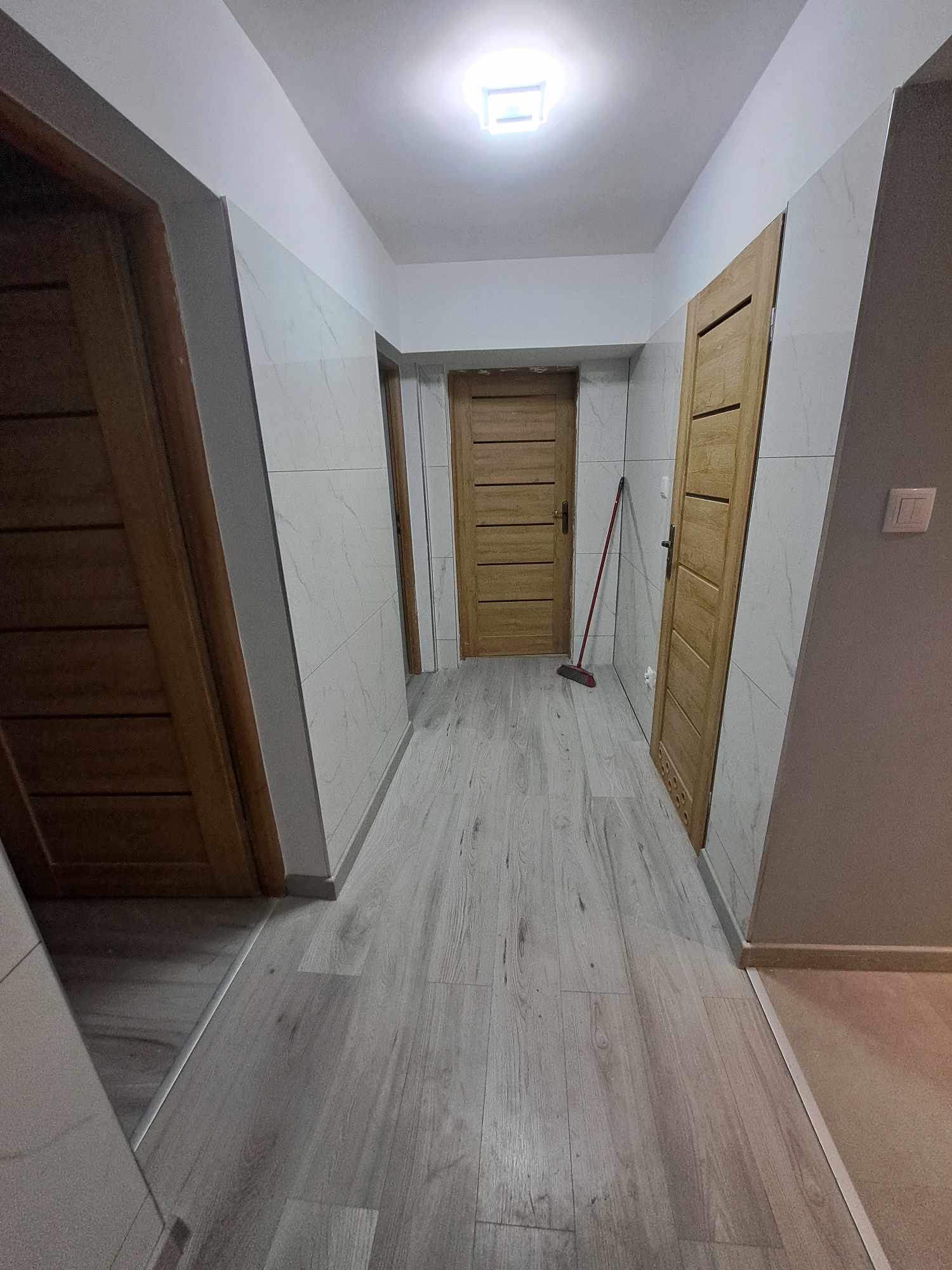 Korytarz po remoncie z panelami podłogowymi imitującymi drewno, wykończony płytkami imitującymi marmur na ścianach, z trzema drewnianymi drzwiami, widoczna czerwona zmiotka oparta o ścianę.