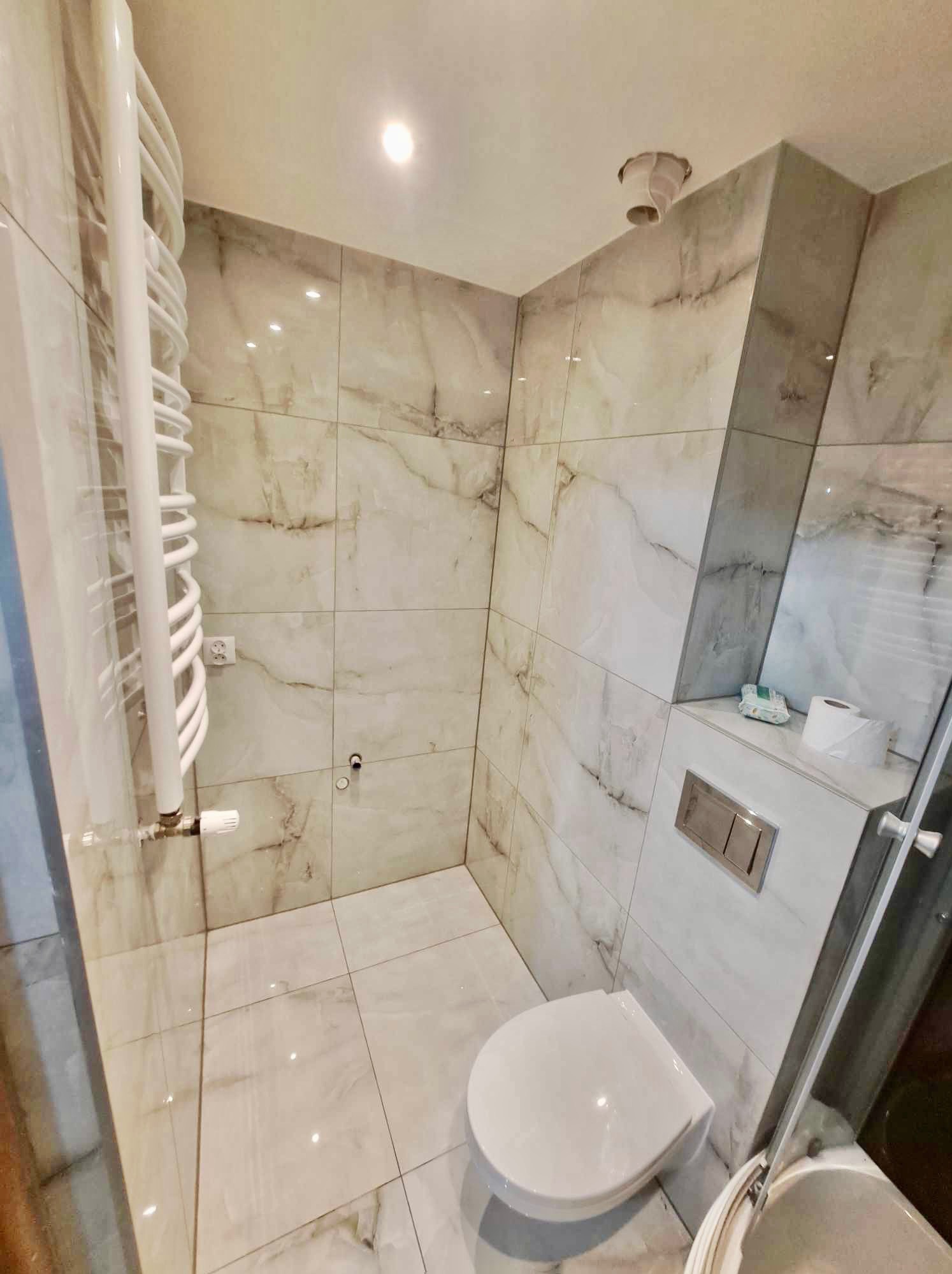 Nowoczesna łazienka z marmurowymi płytkami, podwieszana toaleta z chromowanym przyciskiem spłukującym, biały grzejnik drabinkowy i widoczna wentylacja sufitowa.