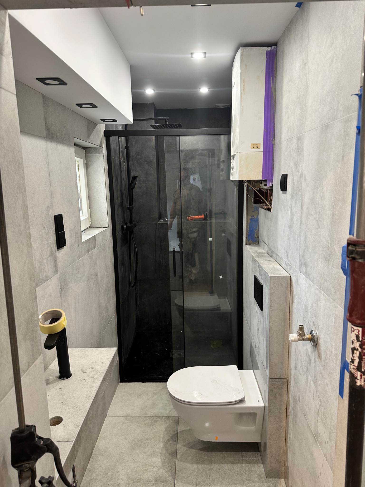 Szara łazienka z czarną kabiną prysznicową typu walk-in, wiszącą toaletą i czarną baterią umywalkową, widoczny bojler zabezpieczony taśmą malarską i narzędzia instalacyjne.