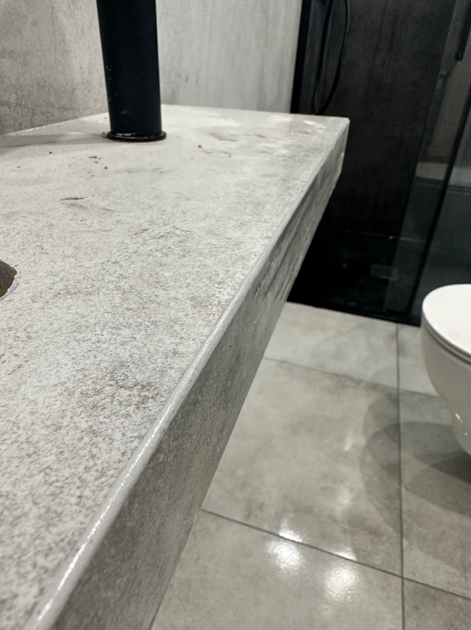 Szary, kamienny blat łazienkowy z czarną armaturą, widok z góry na krawędź blatu i fragment umywalki oraz płytki podłogowe w tle.