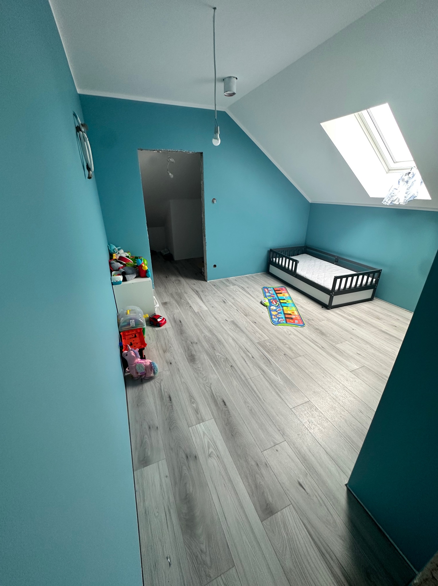 Pokój dziecięcy na poddaszu po remoncie: turkusowe ściany, szare panele podłogowe, dziecięce zabawki i łóżeczko pod oknem dachowym, widoczne niedokończone obramowanie drzwi.