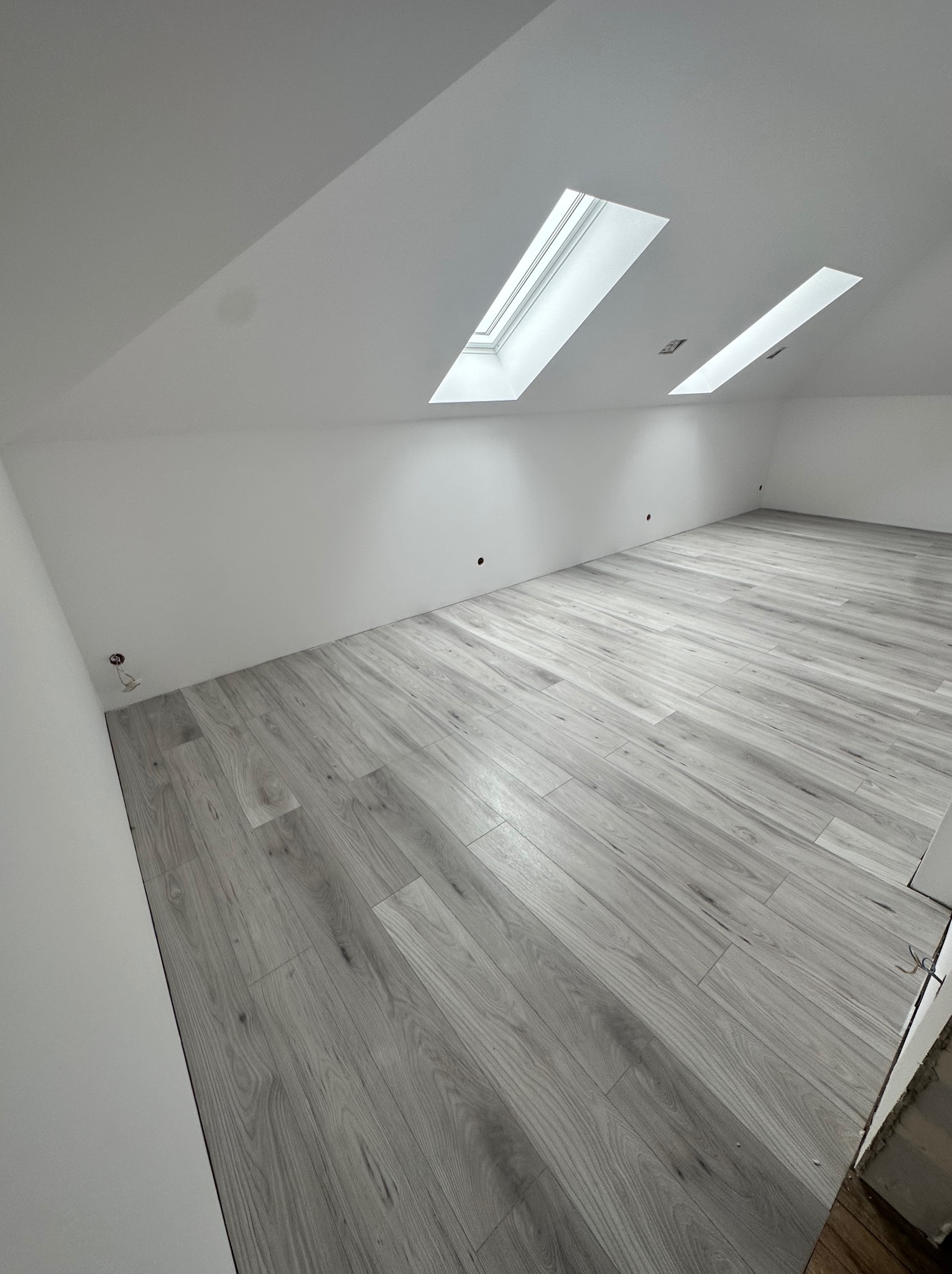 Poddasze z szarymi panelami podłogowymi ułożonymi w jodełkę, dwa okna dachowe w białej ramie, białe ściany z wystającymi przewodami elektrycznymi.