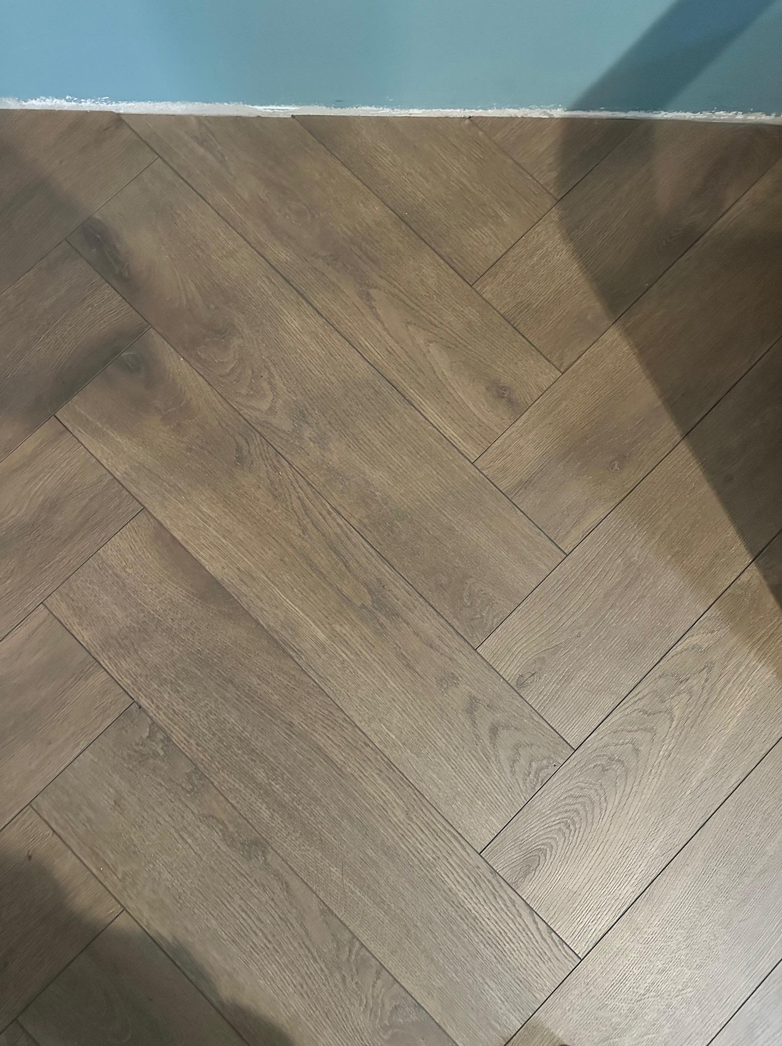 Ułożona pod kątem brązowa podłoga panelowa w jodełkę, widoczna niebieska ściana z białą listwą przypodłogową.