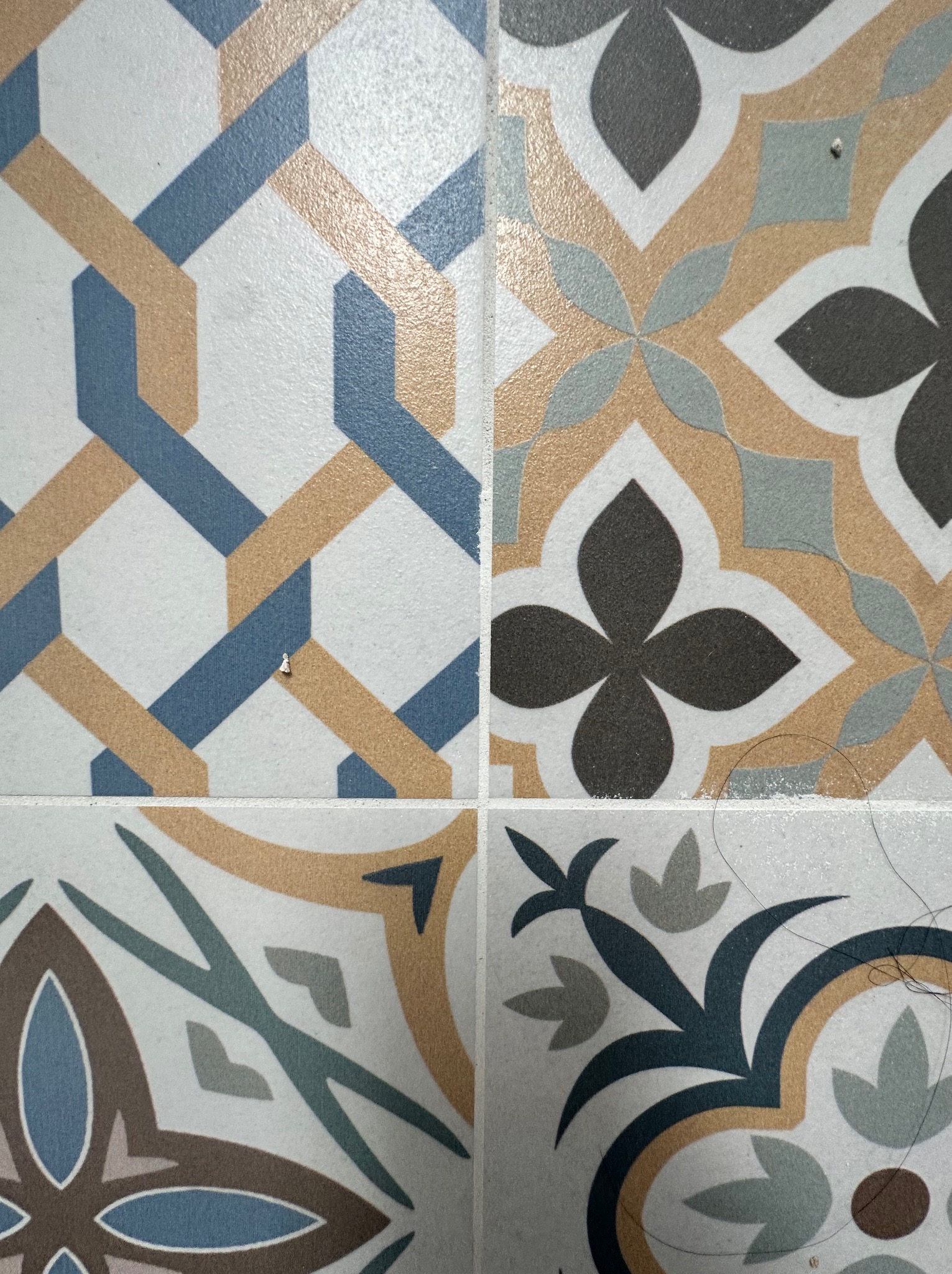 Cztery płytki ceramiczne z różnymi wzorami geometrycznymi i kwiatowymi w odcieniach błękitu, szarości i beżu, z drobnymi zabrudzeniami na powierzchni.