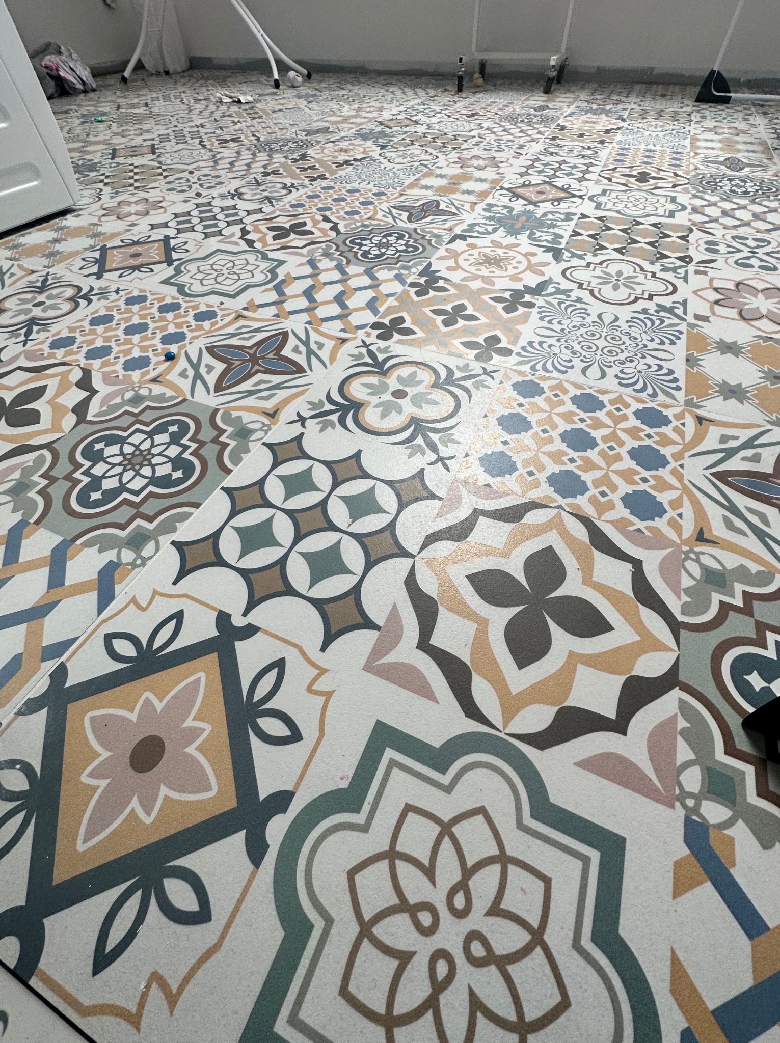 Wzór podłogi z płytek ceramicznych o różnorodnych, geometrycznych i kwiatowych motywach w odcieniach beżu, brązu, szarości i błękitu, ułożonych na jasnej spoinie, widoczne narzędzia malarskie w tle.